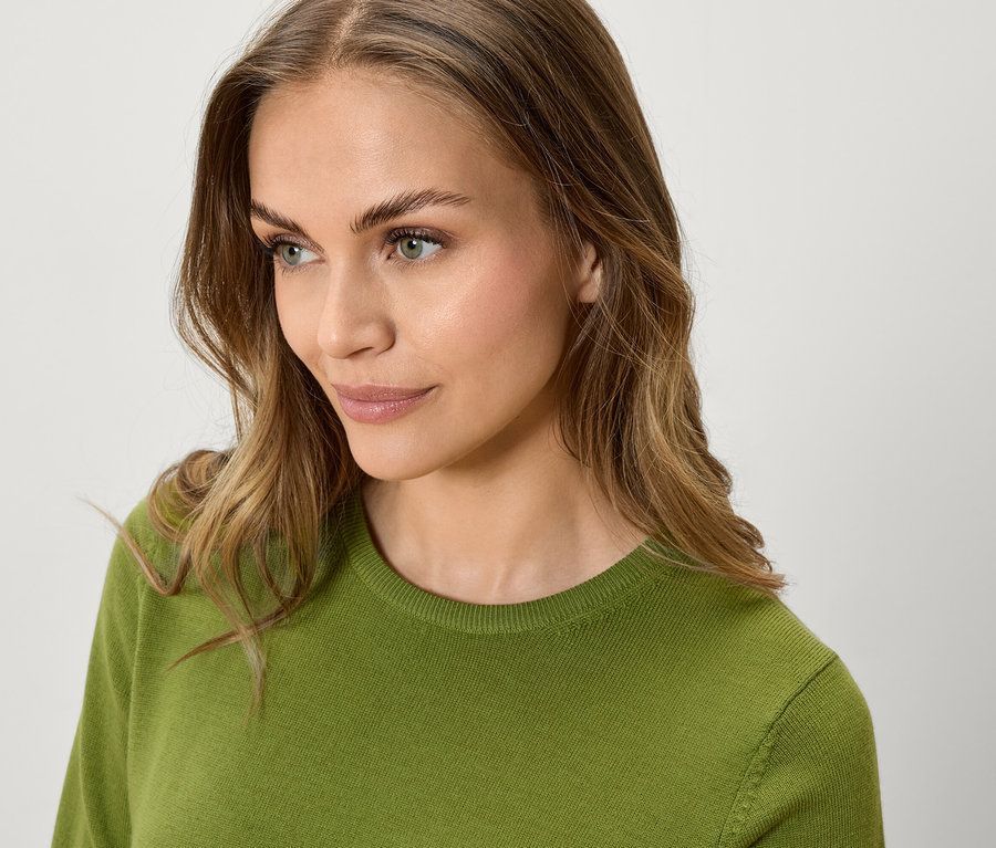 Porträt einer Frau mit gewelltem, braunem Haar und einem grünen Pullover, die zur Seite schaut.