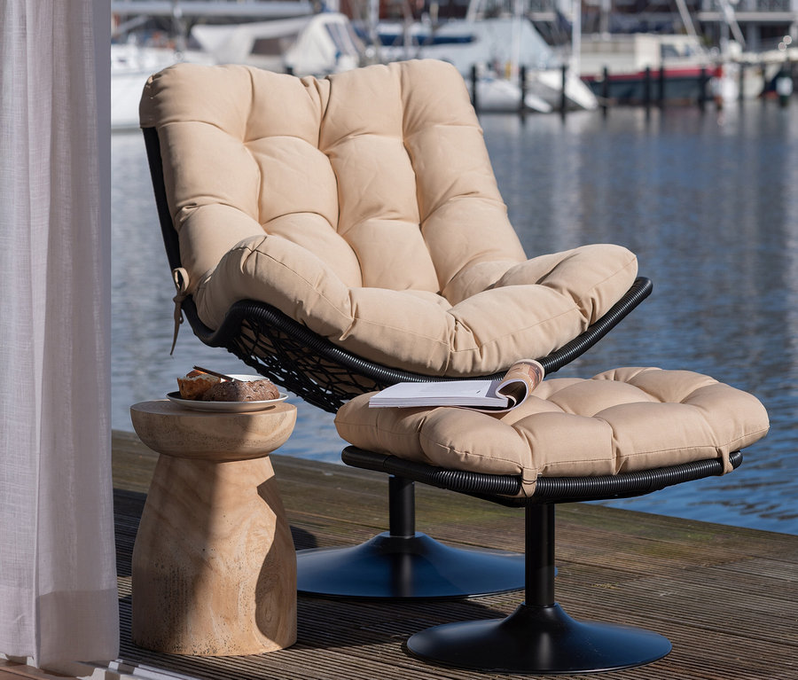 Gepolsterter Sessel mit Hocker und ein naturfarbener Woood Hocker »Bink« aus Holz mit Essen im Freien am Wasser.