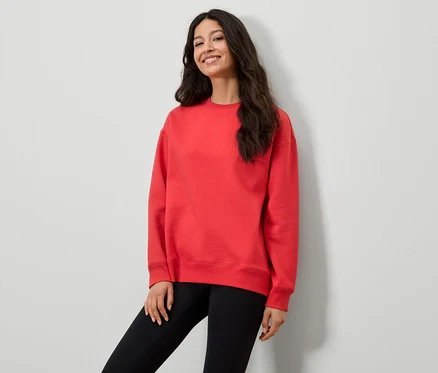 Frau in rotem Sweatshirt und schwarzen Leggings posiert vor einer weißen Wand.