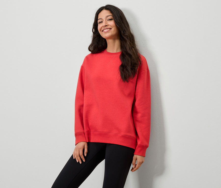 Frau in rotem Sweatshirt und schwarzen Leggings posiert vor einer weißen Wand.