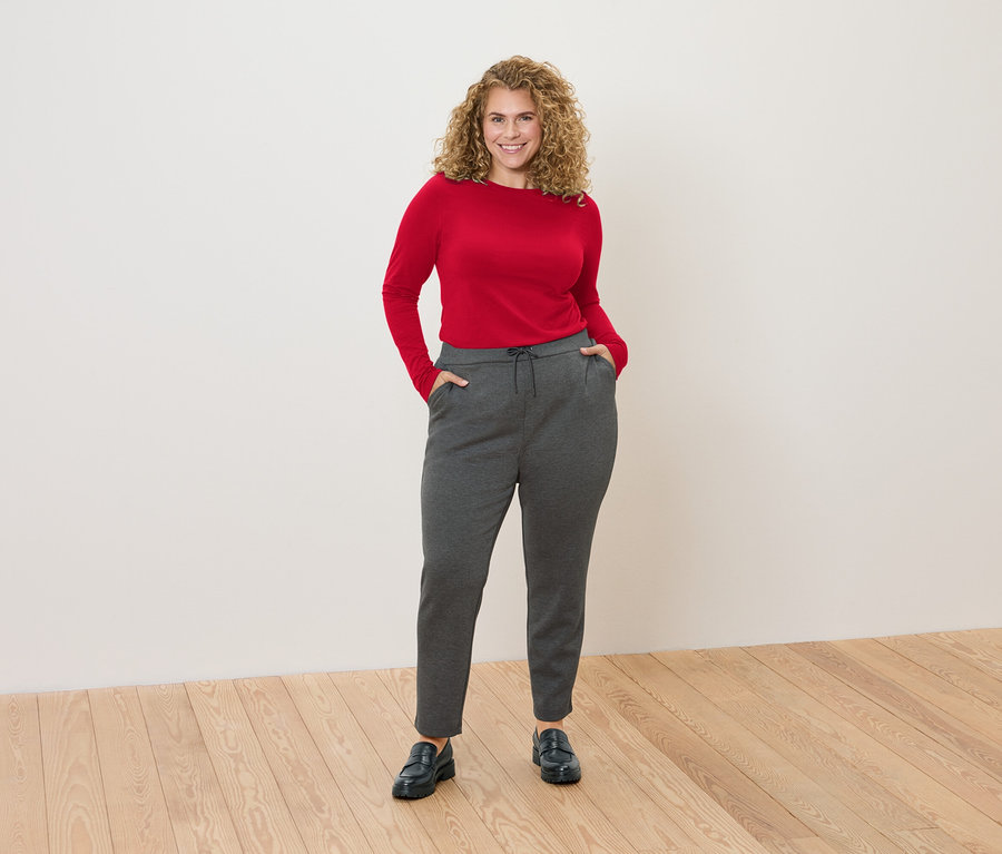 Eine Frau mit lockigem Haar steht vor einer weißen Wand und trägt ein rotes Langarmshirt und graue Jogginghose.