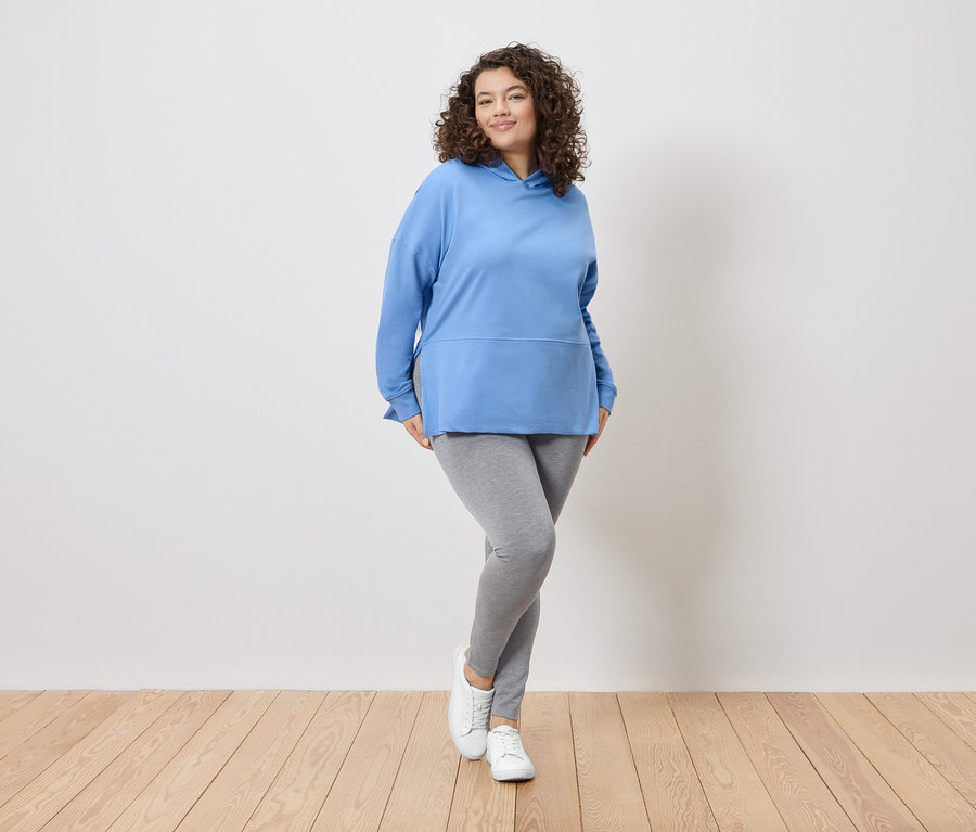 Eine Frau mit lockigem Haar steht vor einer weißen Wand und trägt einen blauen Kapuzenpullover, graue Leggings und weiße Turnschuhe.