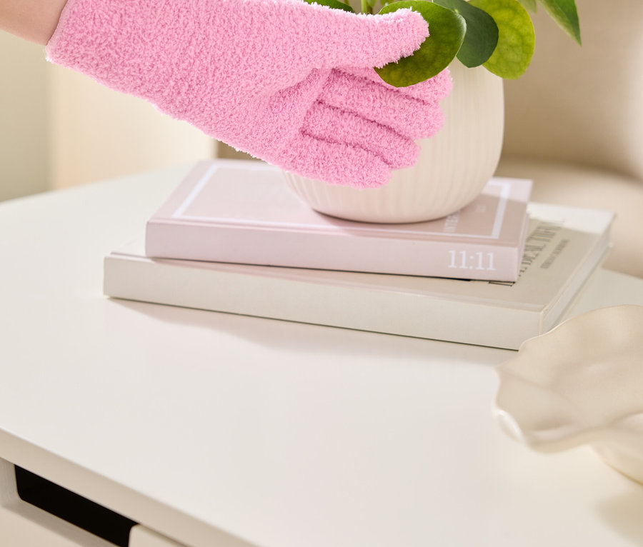 Eine Hand in einem rosa, flauschigen Handschuh berührt die Blätter einer Zimmerpflanze in einem weißen Topf, der auf zwei Büchern auf einem weißen Tisch steht.