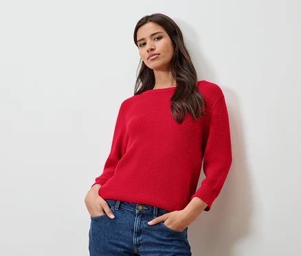 Frau mit rotem Feinstrickpullover und Jeans, die vor einer weißen Wand posiert.