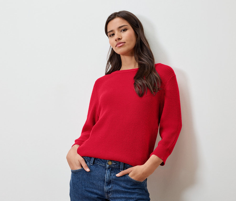 Frau mit rotem Feinstrickpullover und Jeans, die vor einer weißen Wand posiert.