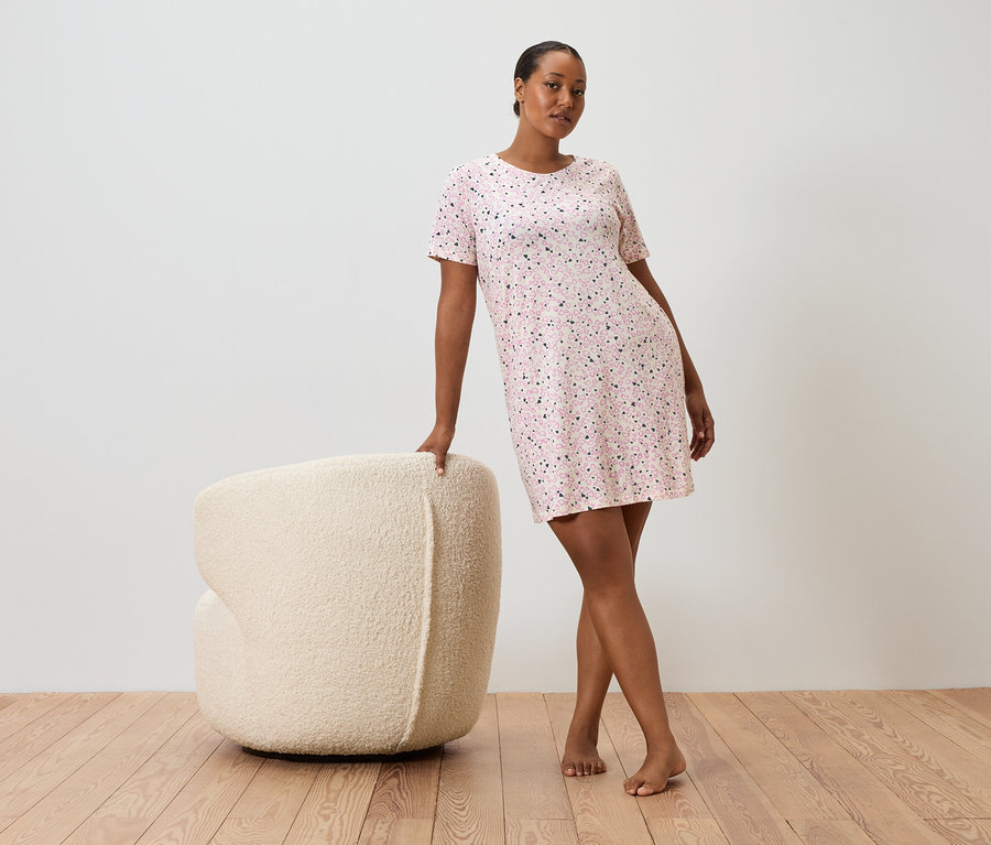 Frau lehnt an einem cremefarbenen Sessel, sie trägt ein kurzärmeliges, rosa gepunktetes Kleid.