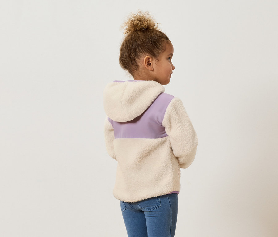 Mädchen mit lockigem Haar, von hinten gesehen, trägt eine Fleecejacke in Beige und Lila und blaue Jeans.