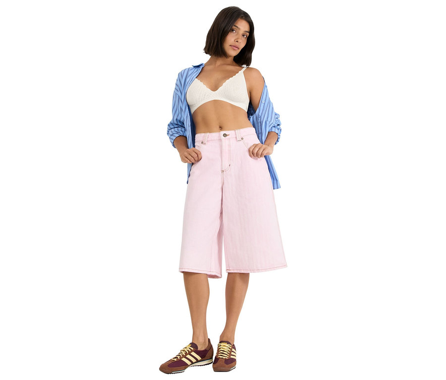 Frau trägt einen weißen Spitzen-BH, eine blaue Hemdbluse und eine rosafarbene Culotte-Hose, dazu braune und gelbe Turnschuhe.