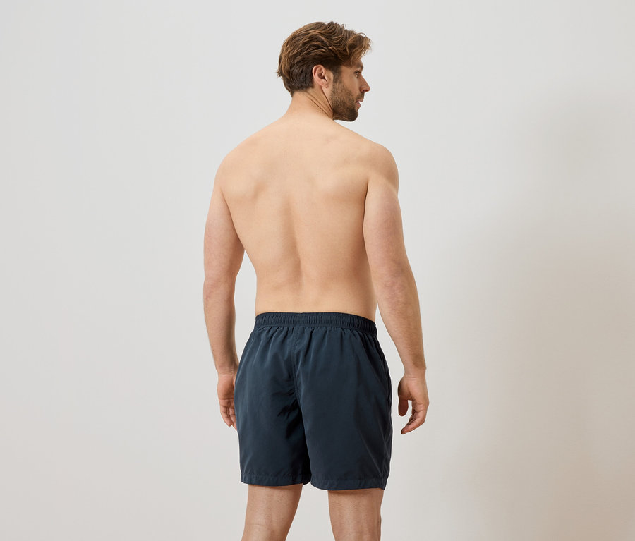 Mann von hinten, mit nacktem Oberkörper und dunkler Badehose vor einem weißen Hintergrund.