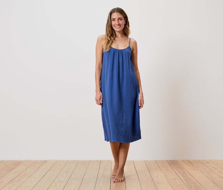 Eine Frau steht barfuß auf einem Holzboden und trägt ein blaues, ärmelloses Kleid.