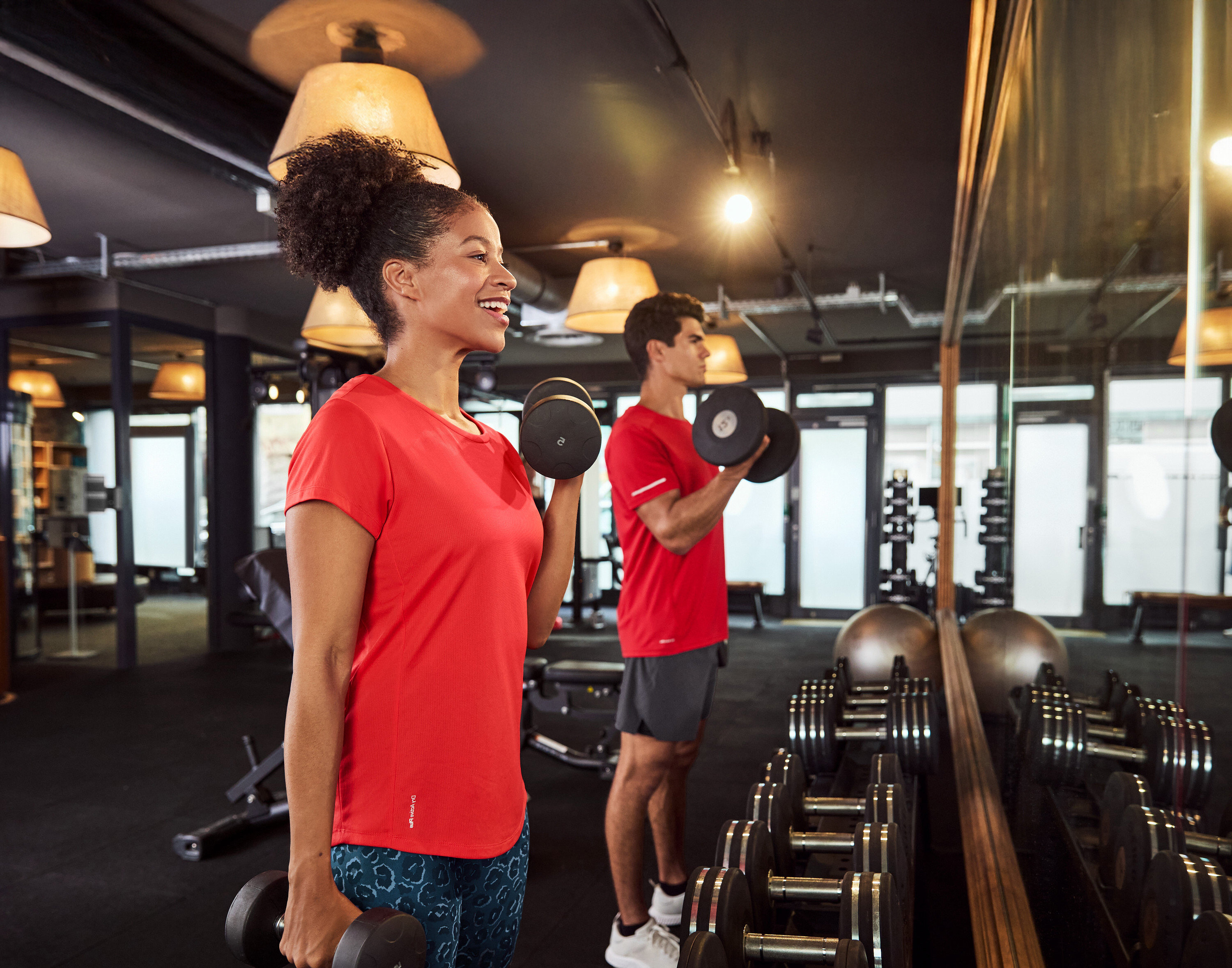 Frau und Mann im Fitnessstudio beim Hanteltraining vor einem Spiegel.