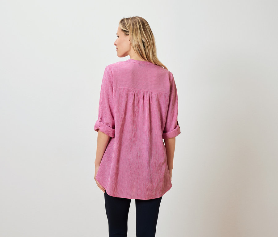 Rückansicht einer Frau mit blonden, schulterlangen Haaren, die eine pinkfarbene Bluse und dunkle Hosen trägt.