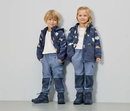 Zwei Kinder stehen nebeneinander und tragen blaue Jacken mit Sonnen-, Wolken- und Regenbogenmotiven, blaue Hosen und passende Schuhe.