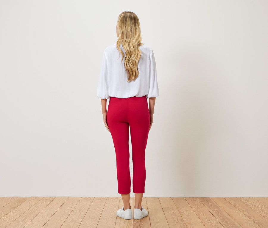Rückansicht einer Frau mit blonden, gewellten Haaren, die eine weiße Bluse und eine rote Hose trägt.