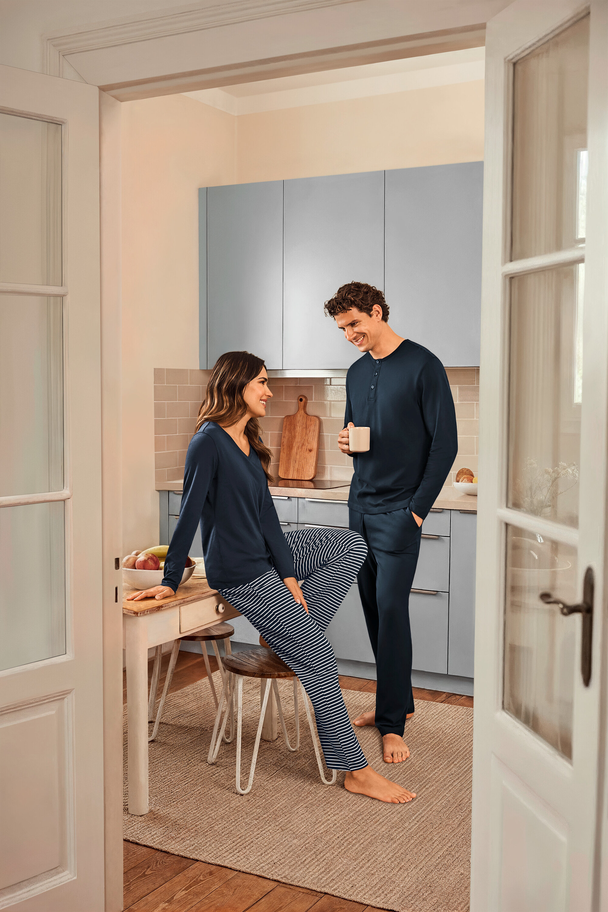 In einer Küche stehen eine Frau, die auf einem Tisch sitzt und einen dunkelblau gestreiften Pyjama und ein dunkelblaues Oberteil trägt, und ein Mann, der eine Tasse hält und einen dunkelblauen Pyjama trägt.