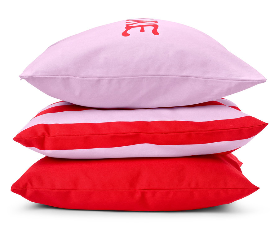 Drei Kissen sind gestapelt: ein rosa Kissen mit Aufschrift, ein rot-weiß gestreiftes Kissen und ein rotes Kissen.