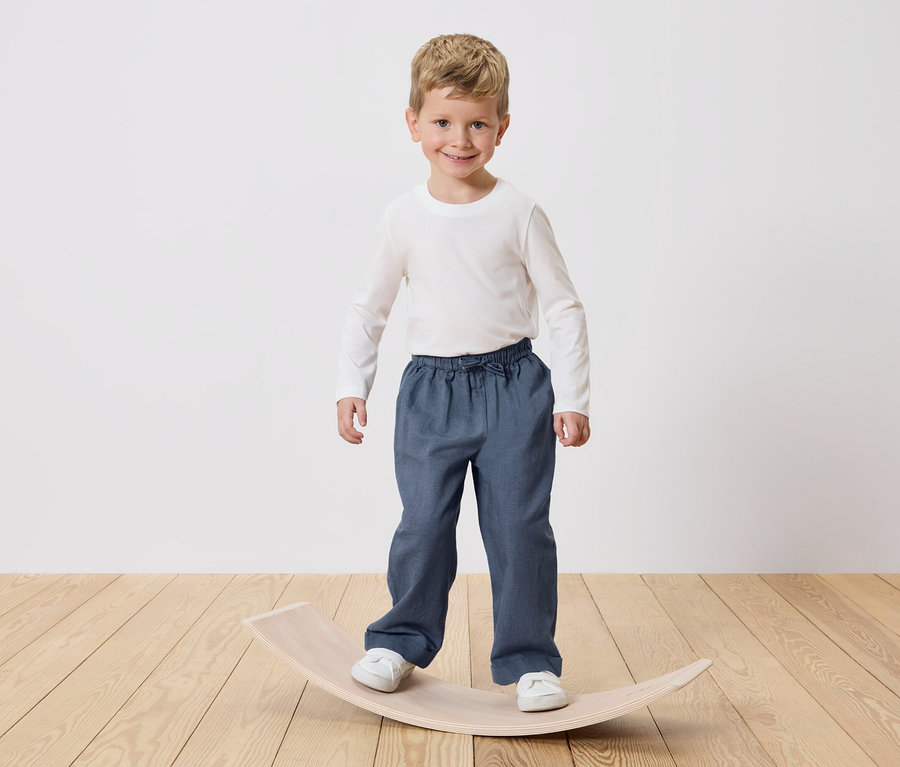 Ein lächelnder Junge mit blonden Haaren steht auf einem hölzernen Balance Board. Er trägt ein weißes Langarmshirt und blaue Hosen.