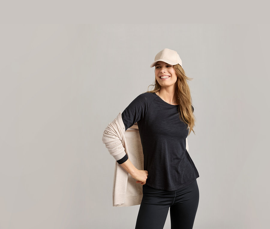 Frau mit beiger Kappe und schwarzem Outdoor-Funktionsshirt. Sie trägt eine beige Strickjacke und schwarze Leggings.