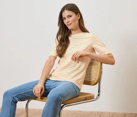 Frau sitzt auf einem Stuhl, trägt ein gelb-weiß gestreiftes T-Shirt und blaue Jeans.