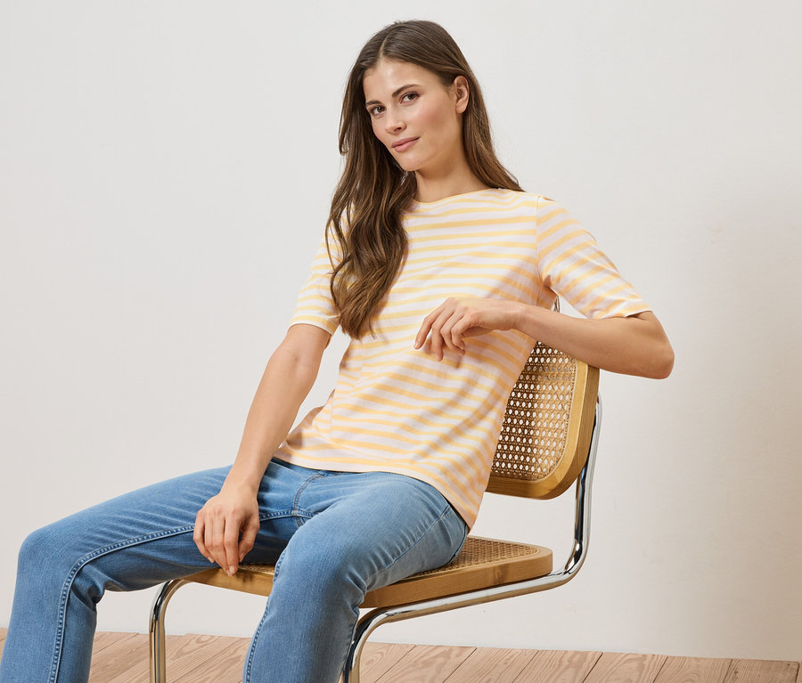 Frau sitzt auf einem Stuhl, trägt ein gelb-weiß gestreiftes T-Shirt und blaue Jeans.