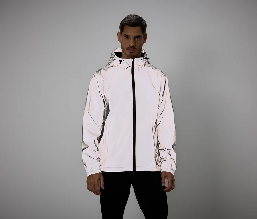 Mann mit grauen Haaren trägt eine reflektierende Jacke mit Kapuze und schwarzem Reißverschluss vor grauem Hintergrund.