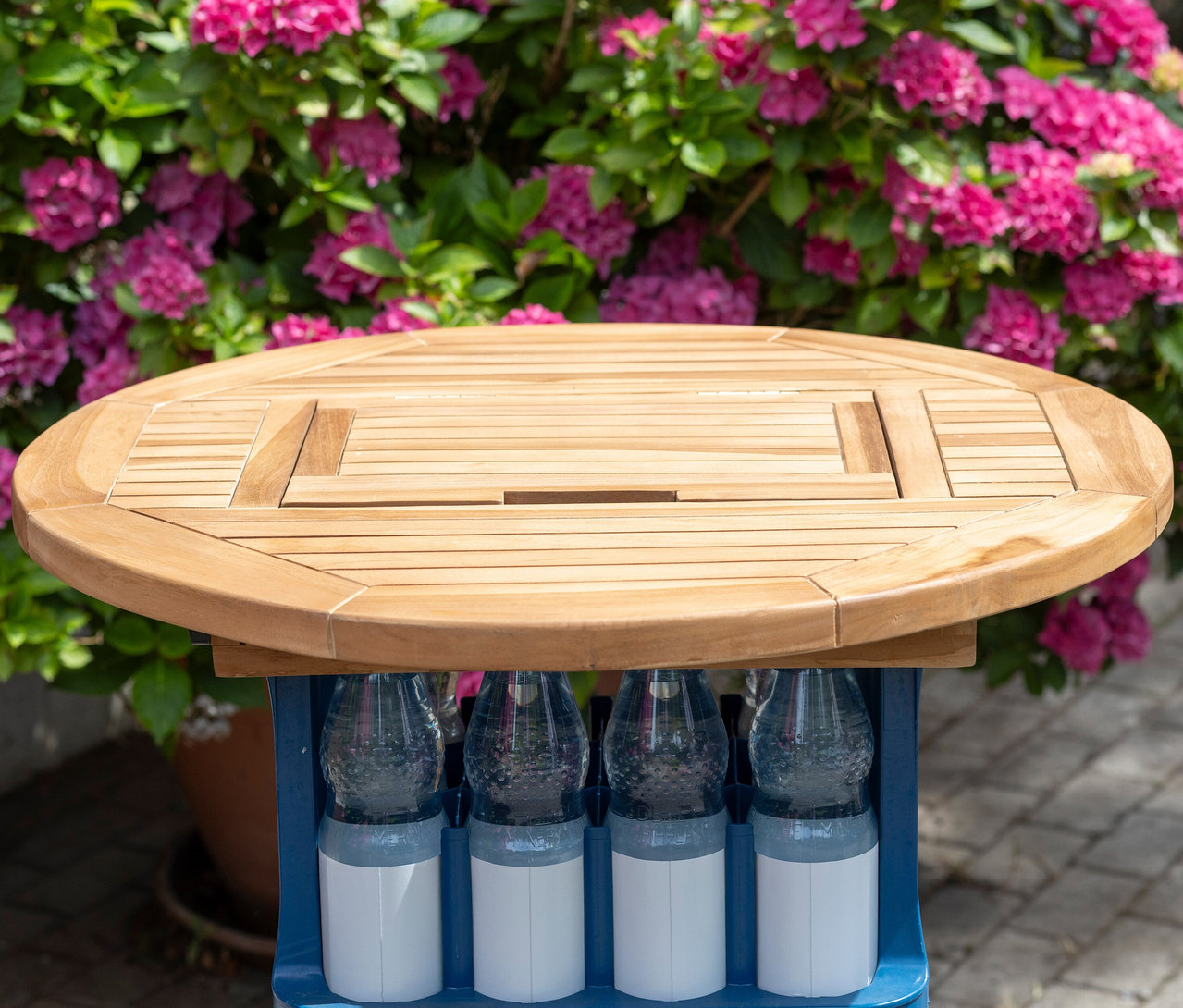 Ein runder Holztisch steht auf einem blauen Getränkekasten, im Hintergrund blühende Hortensien.
