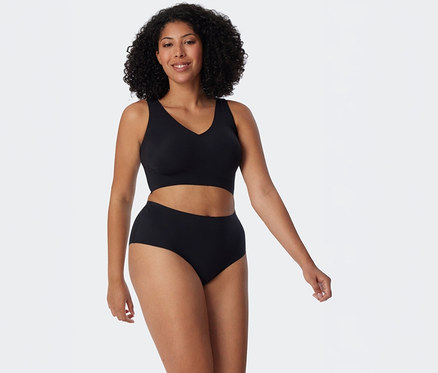 Eine lächelnde Frau mit lockigem dunklem Haar trägt ein schwarzes BH-Oberteil und schwarze SCHIESSER Invisible Slip Maxi vor weißem Hintergrund.