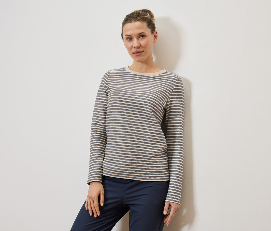 Frau mit Dutt trägt ein grau-weiß gestreiftes Langarmshirt und eine dunkelblaue Hose vor einer weißen Wand.