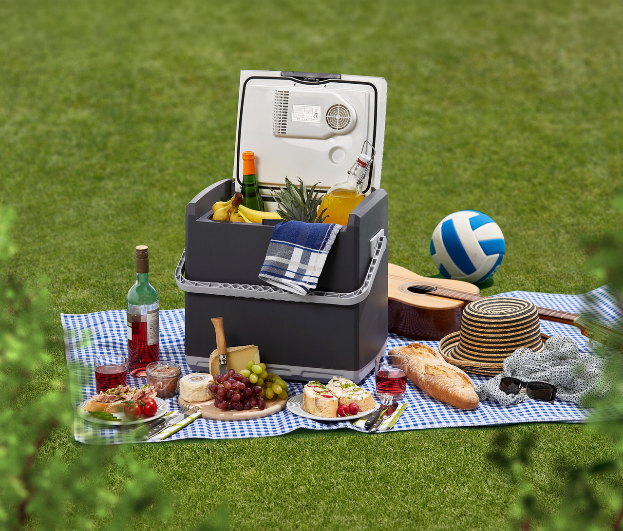 Ein Picknick auf grünem Gras mit einer geöffneten AEG Kühlbox »KK 28«, gefüllt mit Ananas, Bananen und Saft. Auf der blau-weiß karierten Decke liegen Wein, Käse, Brot, Sandwiches, eine Gitarre und ein Volleyball.