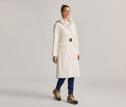 Frau in einem langen, cremefarbenen Mantel mit Gürtel, blauen Jeans und braunen Stiefeln.