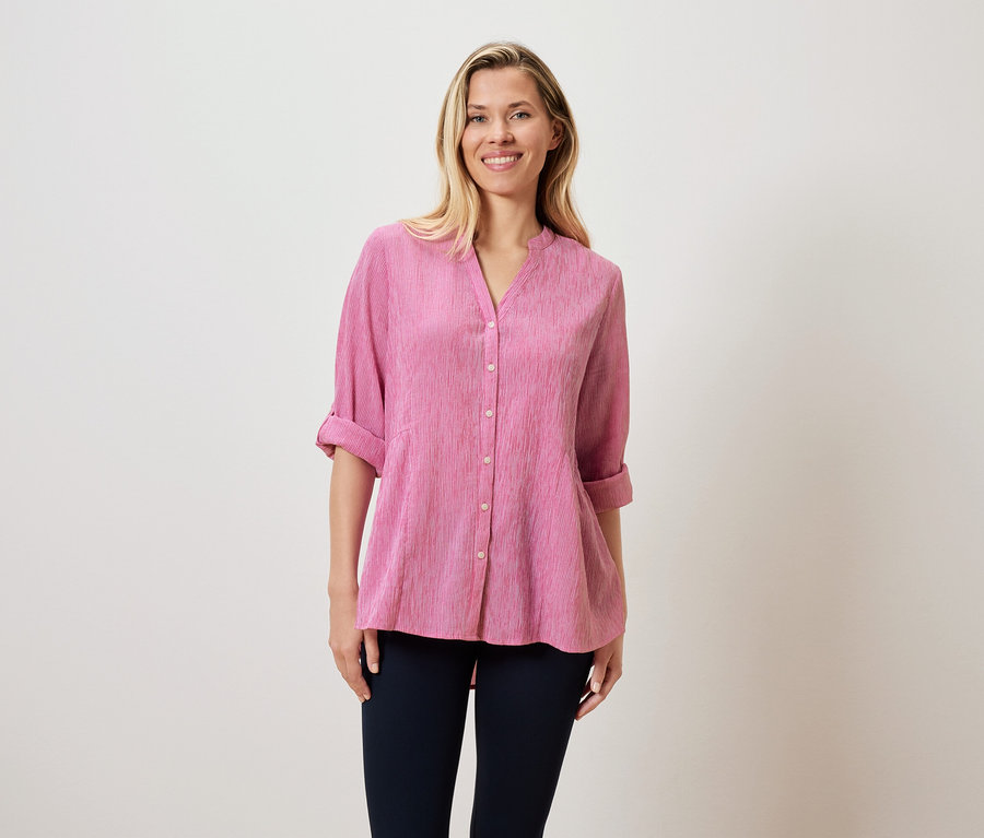 Eine Frau mit blonden Haaren trägt eine pinke Bluse und dunkle Hosen.