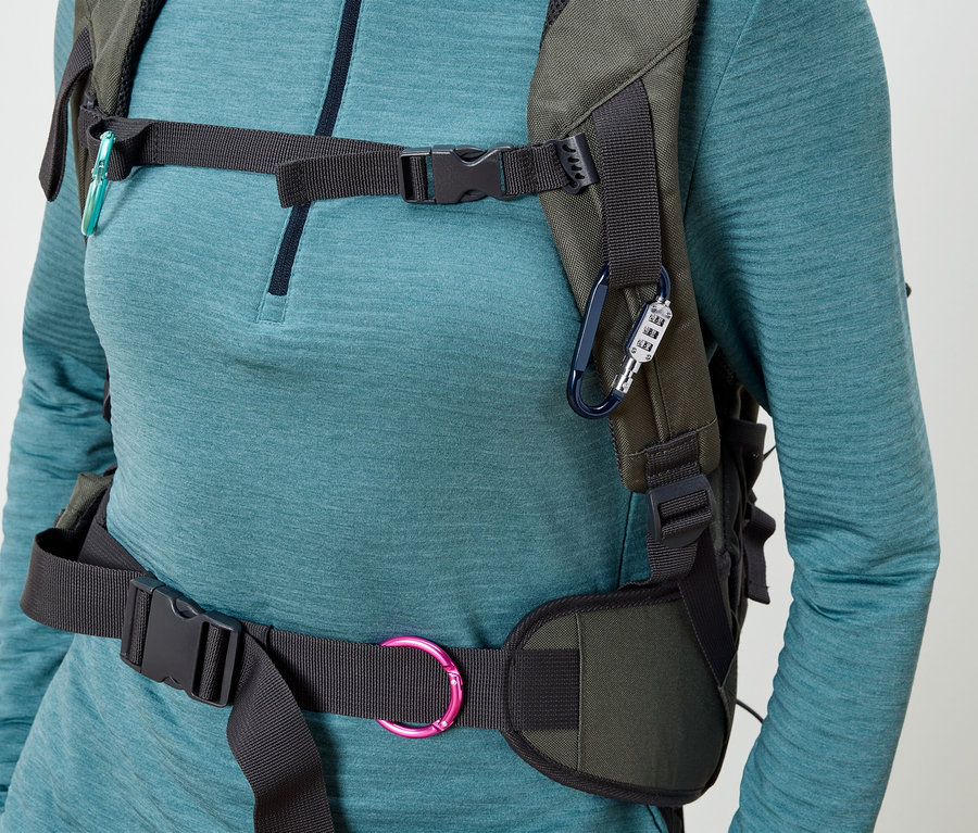 Eine Frau trägt einen olivgrünen Rucksack mit schwarzen Gurten und Schnallen, befestigt mit einem blauen Karabiner mit Zahlenschloss und einem türkisfarbenen Karabiner.