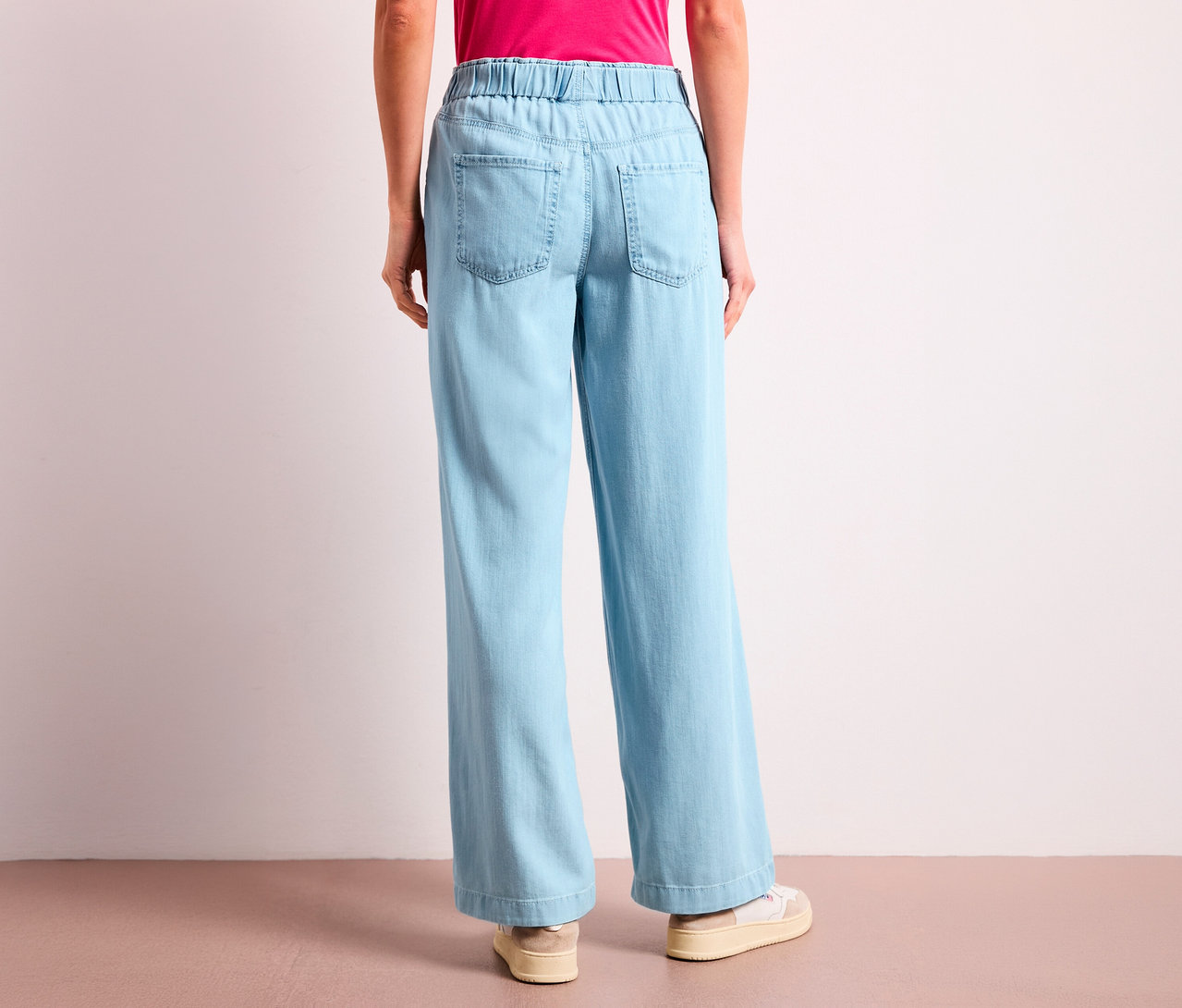 Eine Person von hinten, die hellblaue Street One Jeans »Emee« mit weitem Bein, elastischem Bund und Gesäßtaschen trägt. Dazu ein pinkes Oberteil und helle Sneaker.