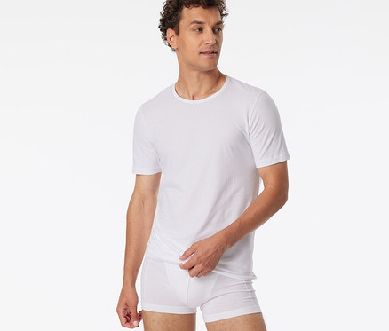 Mann mit lockigem Haar trägt ein weißes SCHIESSER T-Shirt und weiße Boxershorts, blickt nach rechts und zieht den Saum seines T-Shirts hoch.