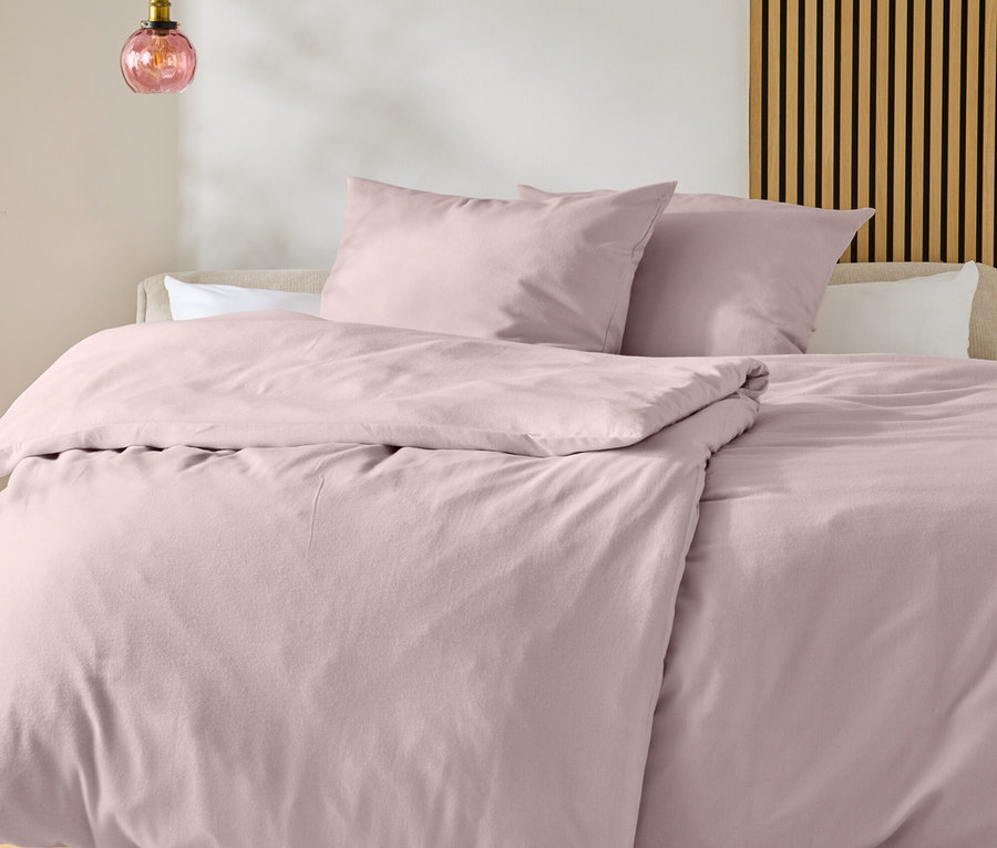 Ein Bett mit hellrosa Bettwäsche und zwei Kissen, davor ein weißer Teppich mit schwarzem Muster auf Holzboden. Eine rosa Glas-Pendelleuchte hängt links, eine Holzlattenwand ist rechts.