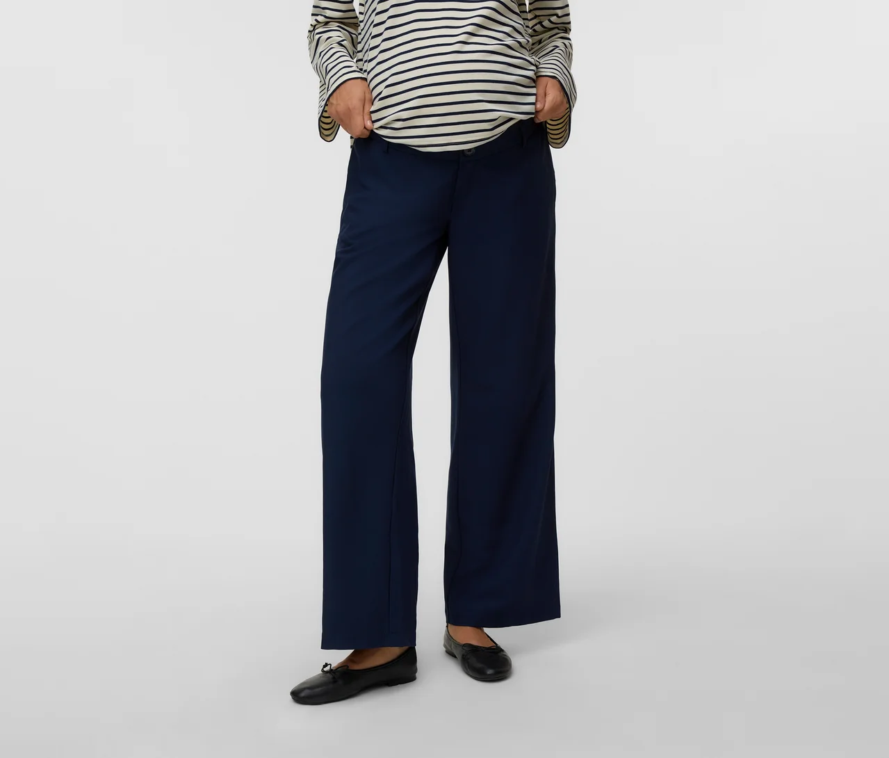 Eine Person trägt ein gestreiftes Langarmshirt, eine marineblaue weite Umstandshose und schwarze Ballerinas und passt den Hosenbund an.