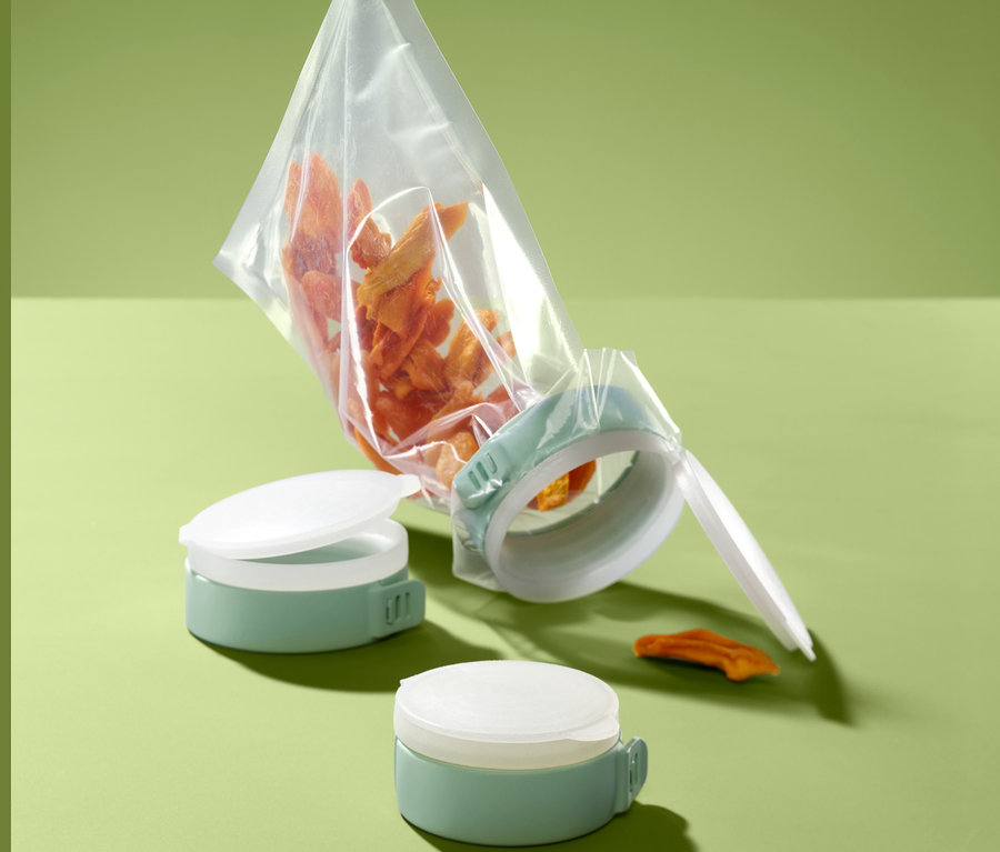 Eine durchsichtige Tüte mit orangefarbenen Essensstücken wird mit 3 Aroma-Tütenverschlüssen in Grün verschlossen. Zwei weitere Verschlüsse liegen daneben.