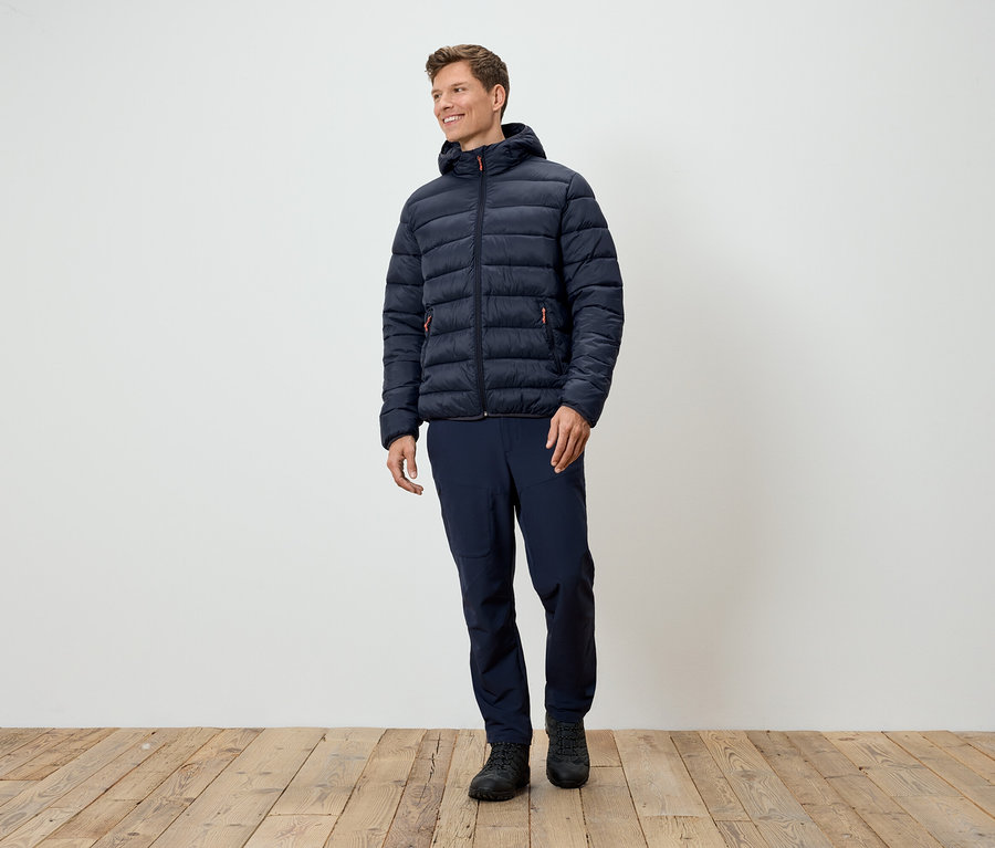 Ein Mann steht und trägt eine dunkelblaue Steppjacke, dunkelblaue Hosen und schwarze Schuhe.