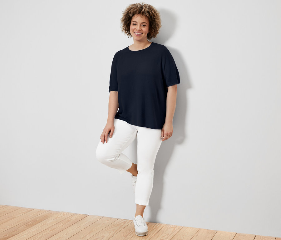 Eine Frau, die sich an eine Wand lehnt, trägt ein marineblaues Feinstrick-Shirt, weiße Hosen und Turnschuhe.