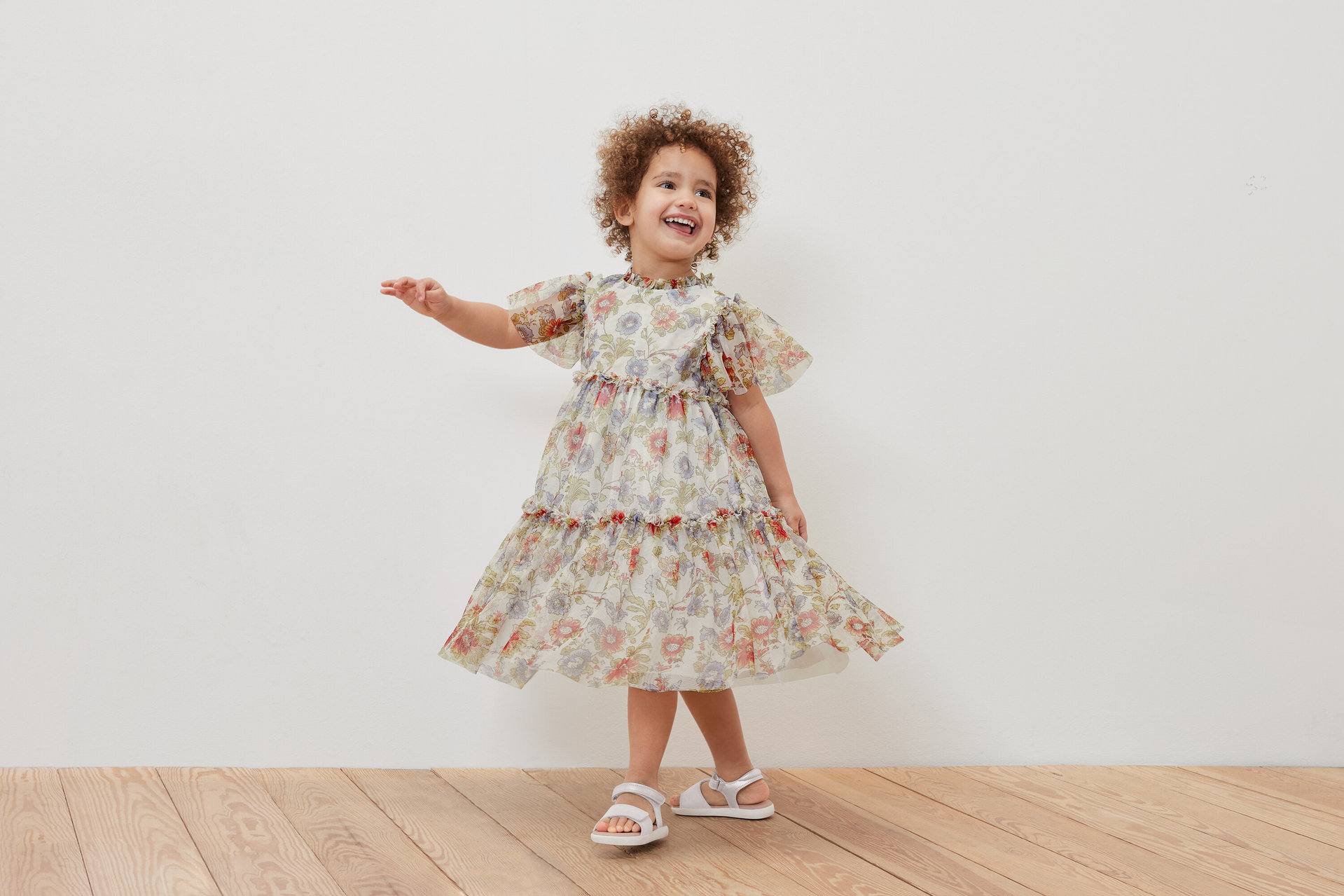 Ein lächelndes Mädchen posiert in einem geblümten Kinder-Tüllkleid und weißen Sandalen.