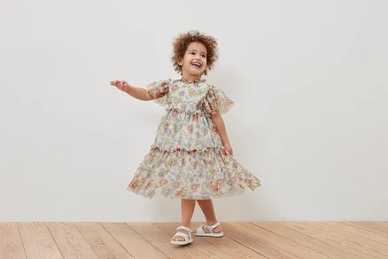 Ein lächelndes Mädchen posiert in einem geblümten Kinder-Tüllkleid und weißen Sandalen.