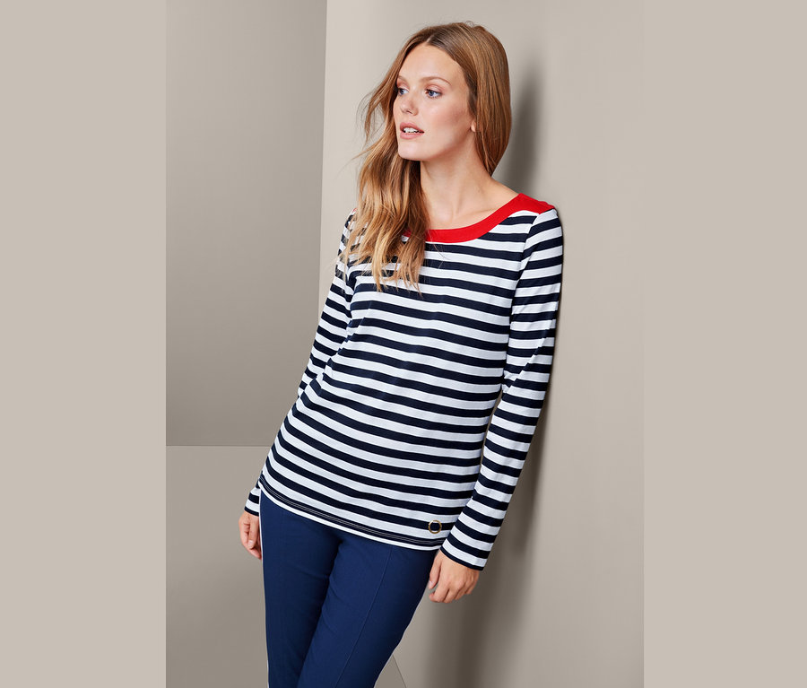 Frau lehnt an Wand, trägt gestreiftes Langarmshirt und blaue Hose.