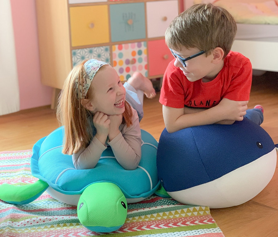 Mädchen liegt auf dem Bauch auf Pool Buddy Schildkröte und lacht Jungen an, der auf Pool Buddy Wal im Kinderzimmer sitzt.