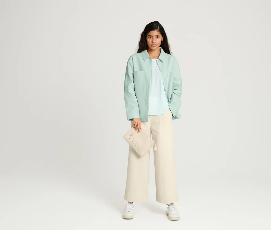 Frau posiert in NAH/STUDIO Culotte aus ungefärbter Baumwolle, NAH/STUDIO Blusenjacke aus recycelter Baumwolle, NAH/STUDIO T-Shirt REFIBRA™ Ocean Blue und NAH/STUDIO Kosmetiktasche aus Baumwolle.
