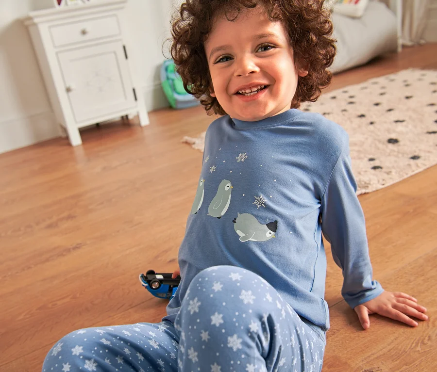 Lächelndes Kind sitzt auf dem Boden und trägt einen Kinder-Glow-in-the-dark-Pyjama mit Pinguinprint.