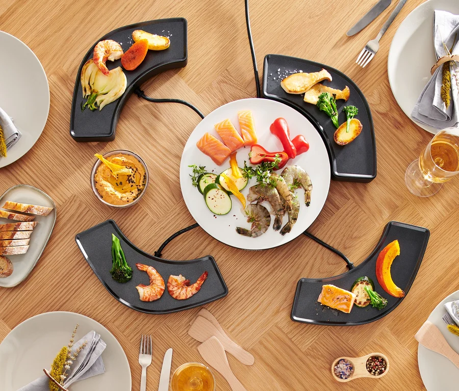 Draufsicht auf einen Tisch mit »Dinner4All« Tischgrill, Tellern, Speisen und Getränken.