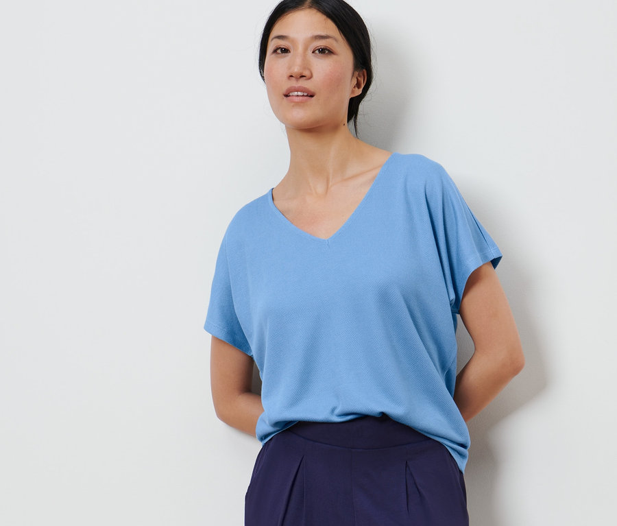 Frau posiert vor einem weißen Hintergrund und trägt ein hellblaues, strukturiertes Shirt und dunkelblaue Hosen.