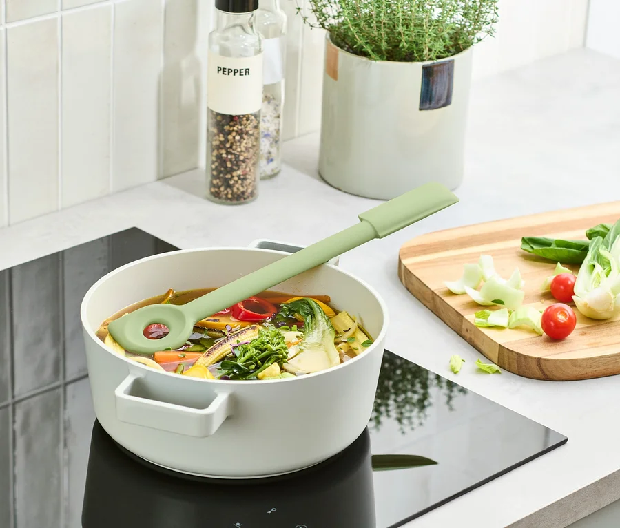Ein grüner Multifunktions-Kochlöffel liegt in einem Topf mit Suppe auf einem Induktionskochfeld.