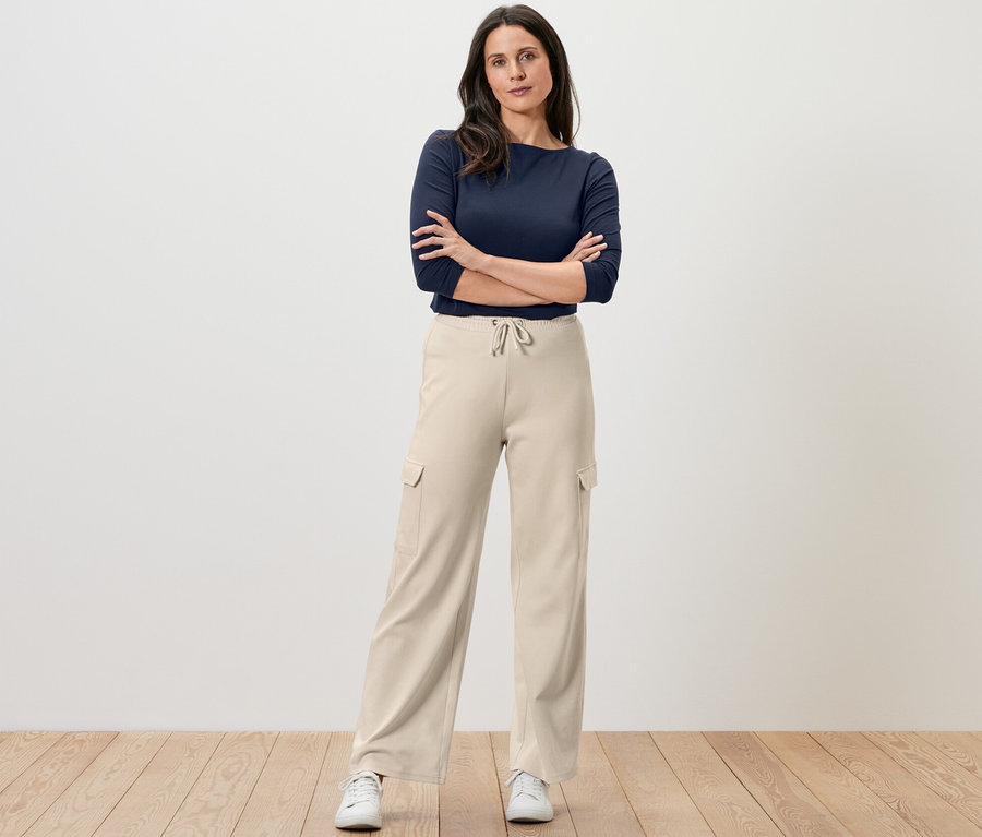 Eine Frau steht mit verschränkten Armen und trägt eine cremeweiße Sweathose mit Cargo-Taschen und ein marineblaues Oberteil.