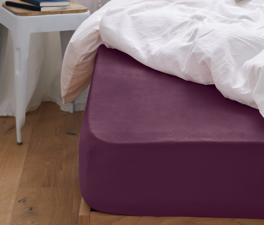 Detailansicht eines Bettes mit einem auberginefarbenen Biber-Spannbettlaken, einer weißen Bettdecke und einem rosa Kissen.